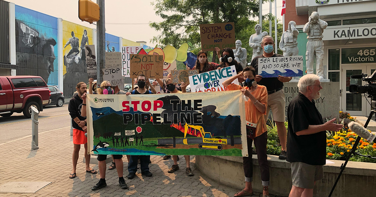 A group of people hold a "Stop the Pipeline" banner and other signs below a smoky sky in front of a local government office in Kamloops.