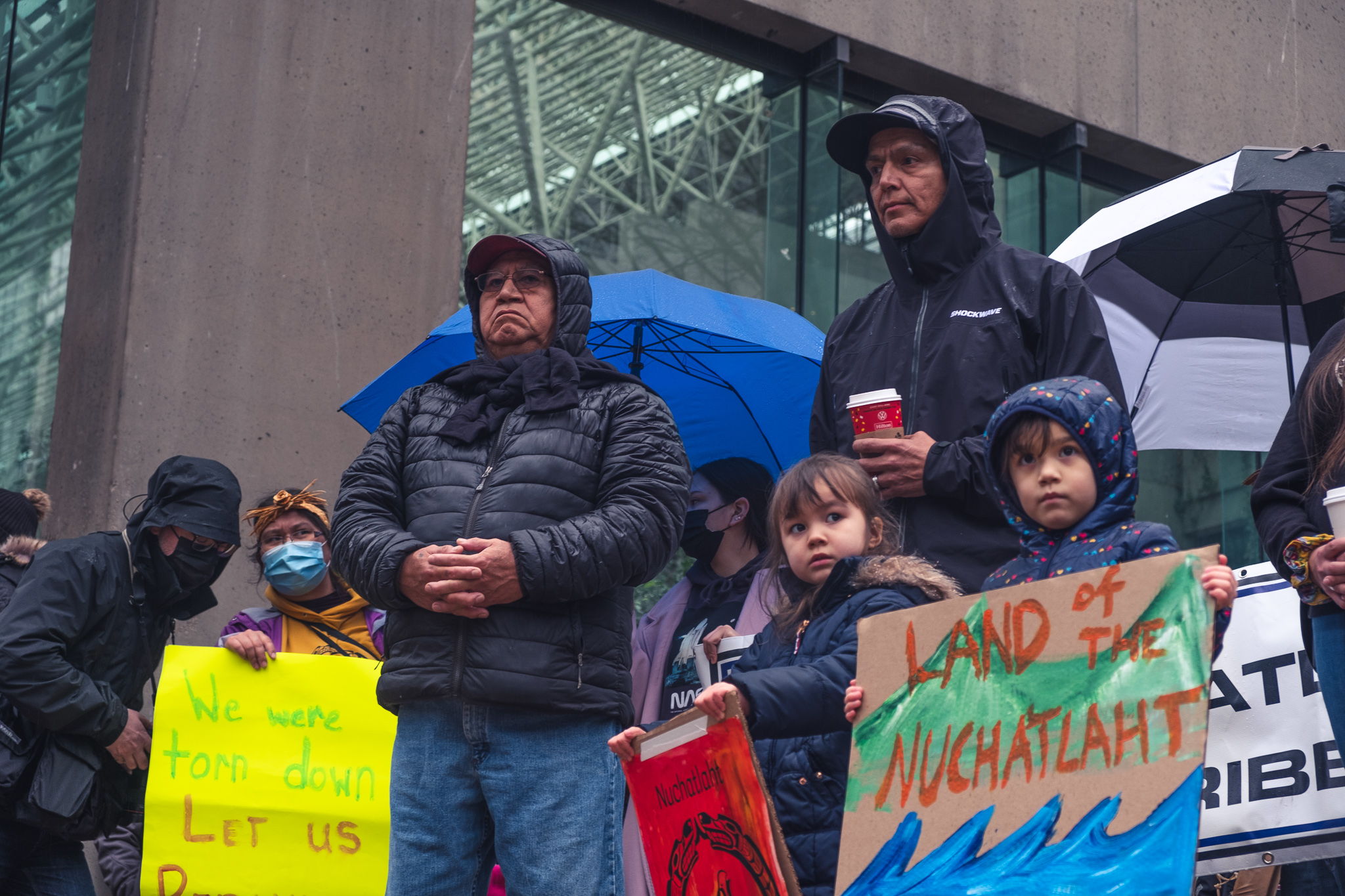 Members of Nuchatlaht Nation stand in the rain holding brightly coloured signs that say "WE WERE TORN DOWN" and "LAND OF THE NUCHATLAHT". End of image description.