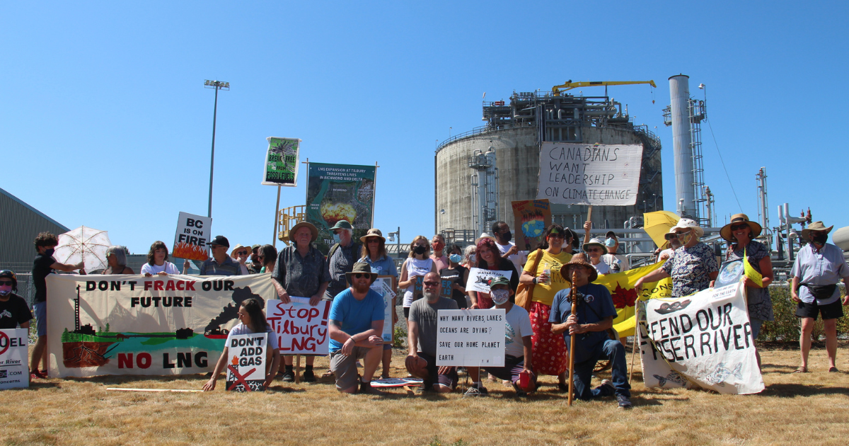 A group of people standing in front of a fracking facility holding various anti-fracking signs. End of image description.