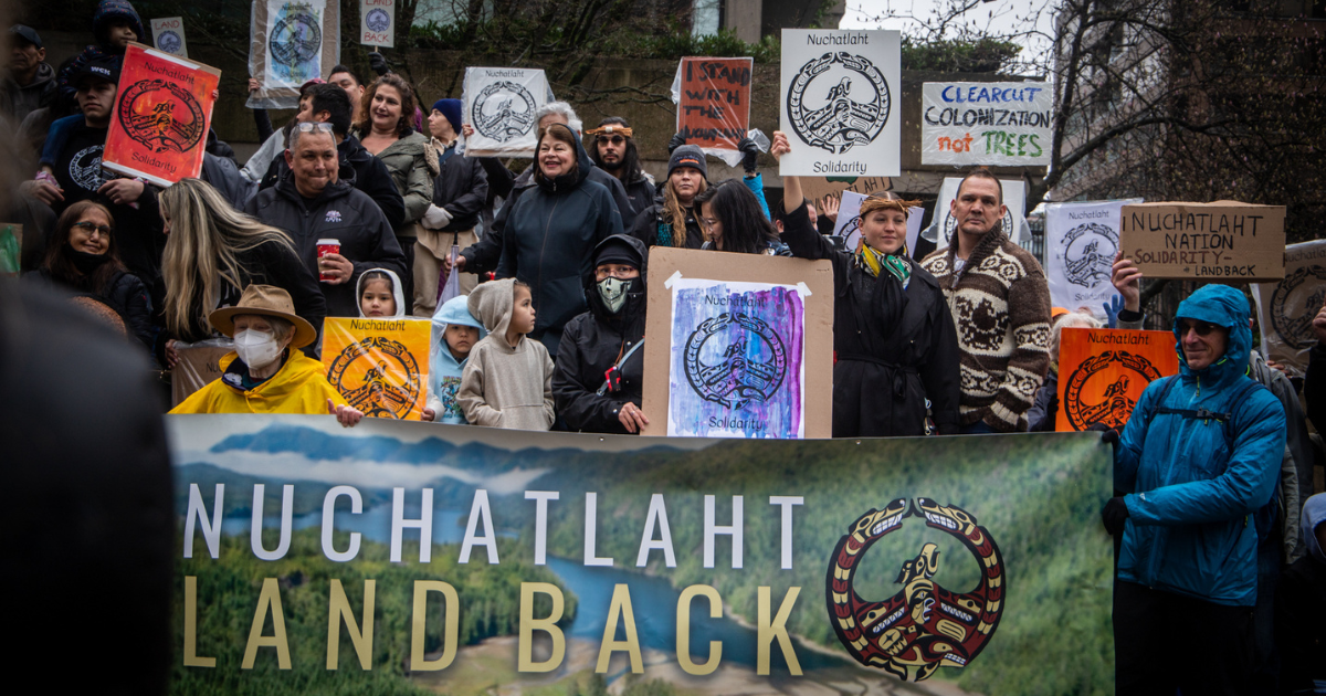 A group of people holding up various signs and banners expressing support for the Nuchatlaht Nation. End of image description. 