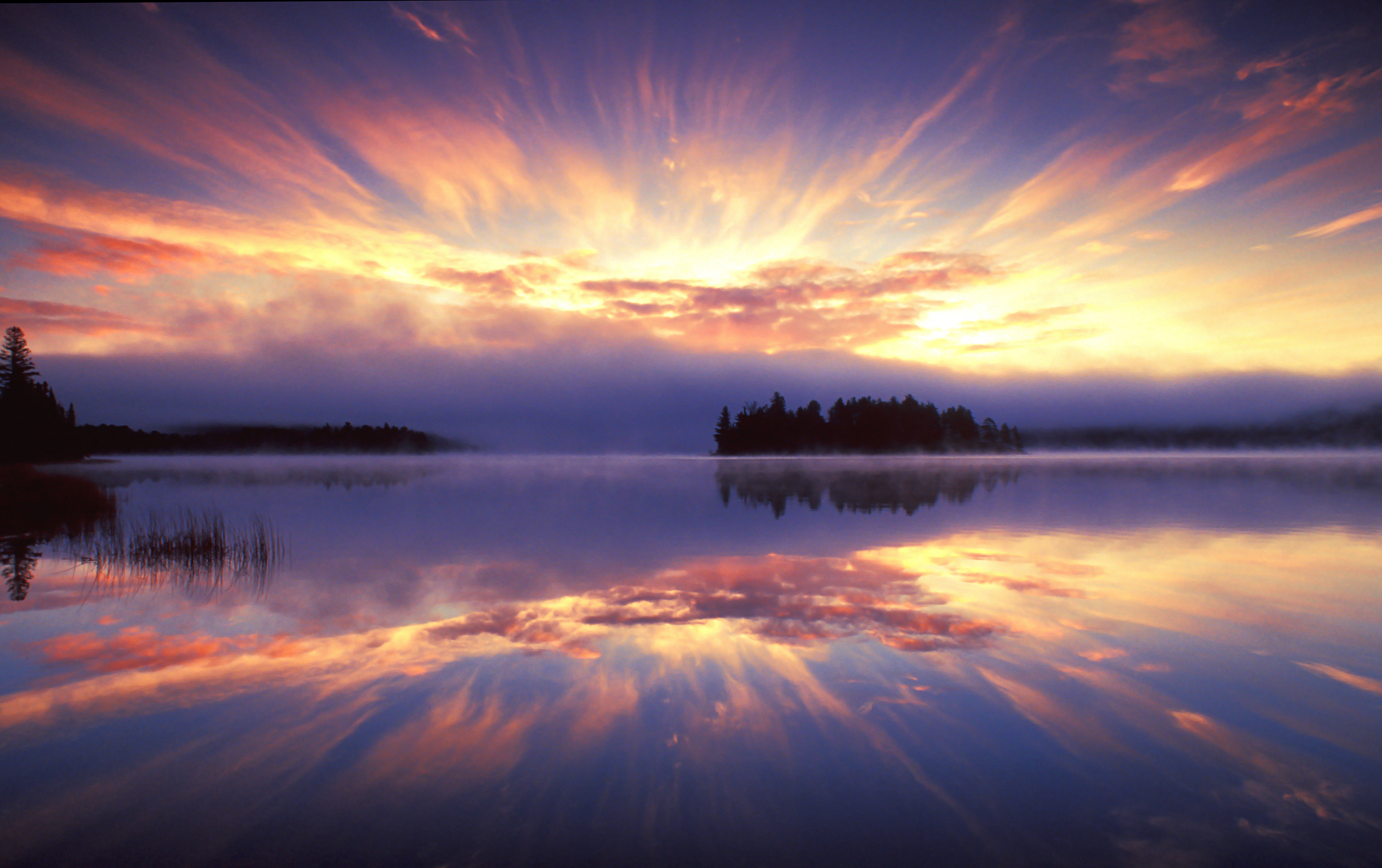 A bright sunburst streaks out from behind pink clouds on a periwinkle sky. Across the lake on a distant shore the forests are silhouetted in black. The water of the lake reflects the sky like a mirror.