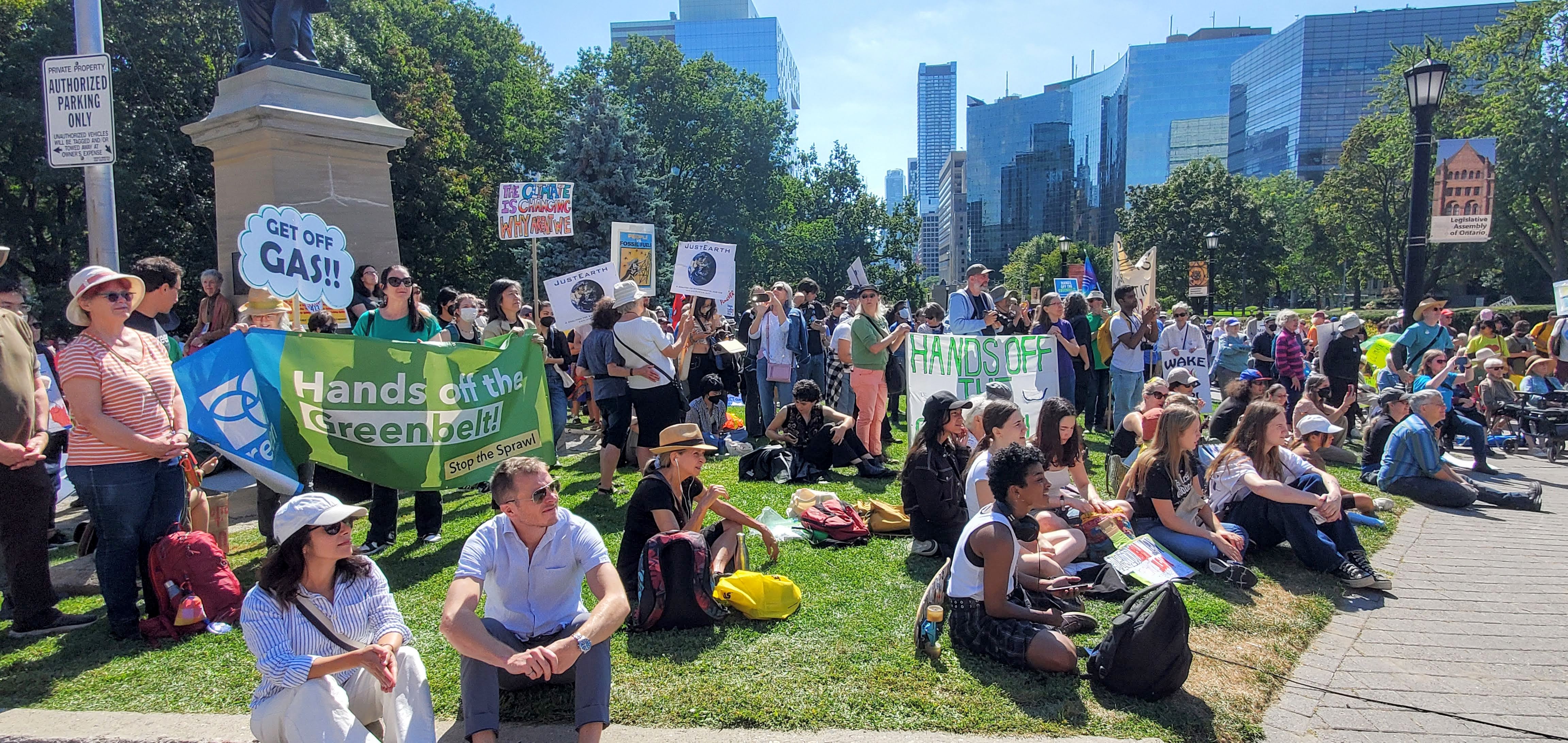A large crowd gathers on the grass in front of Queen's park. Many hold signs and banners, including one shaped like a grey cloud that says