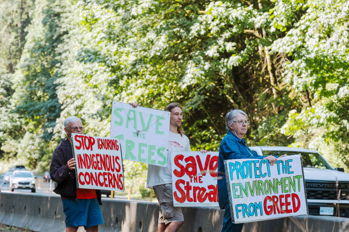Supporters holding home-made signs reading "Protect the environment from greed" "Save the stream" "Stop ignoring indigenous concerns" "Save the trees". 