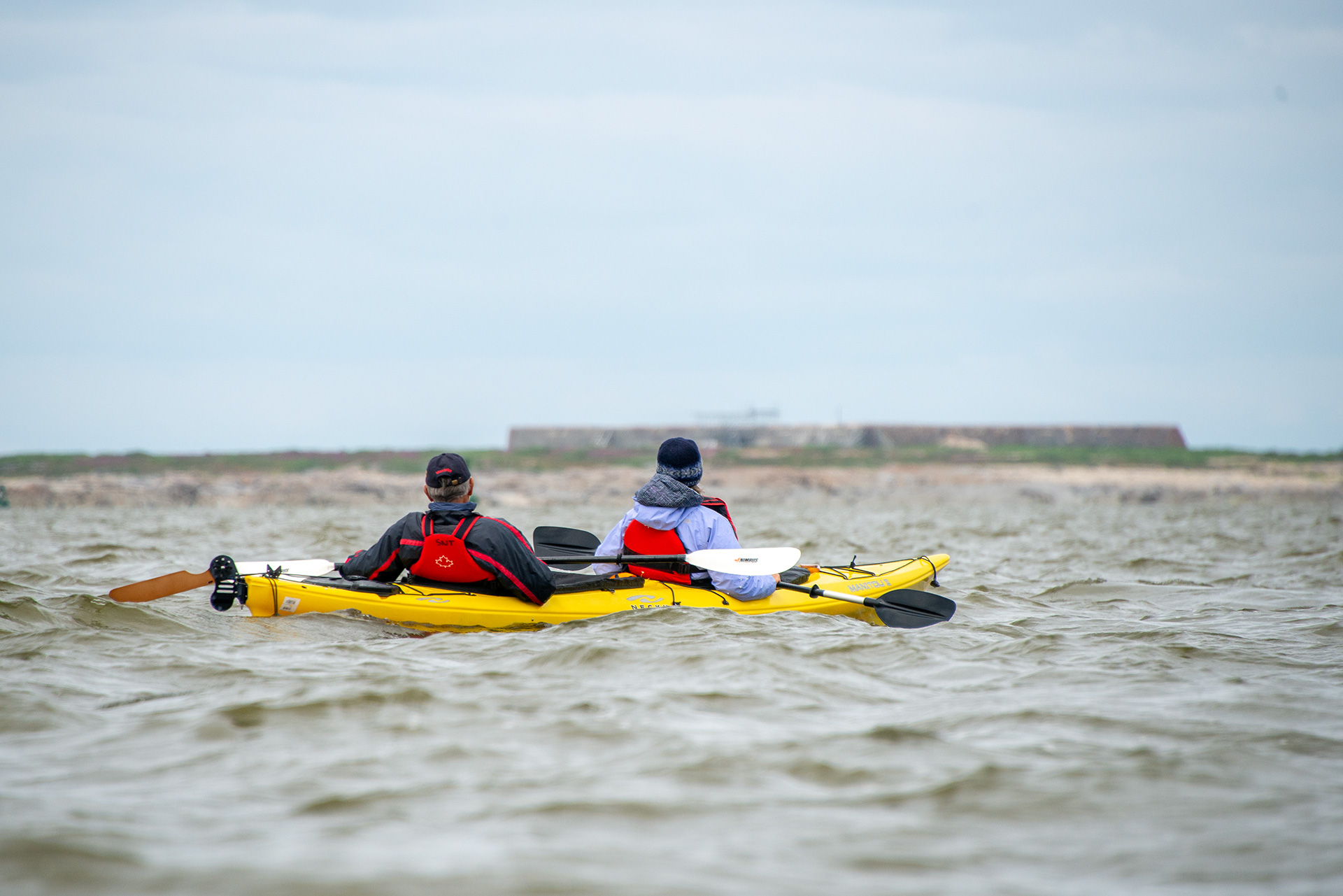 two people in winter gear sit in a bright yellow kayak as it floats on the cold grey water of Hudson's Bay.
