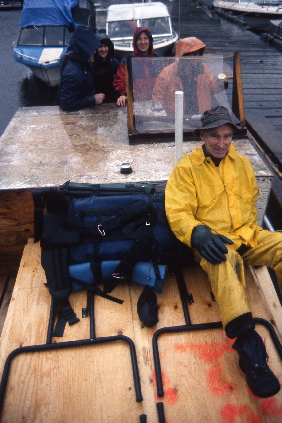 Don Gillespie in a yellow rainsuit on the front of a boat tied to a dock. The captain and a few other people and boats are in the background.