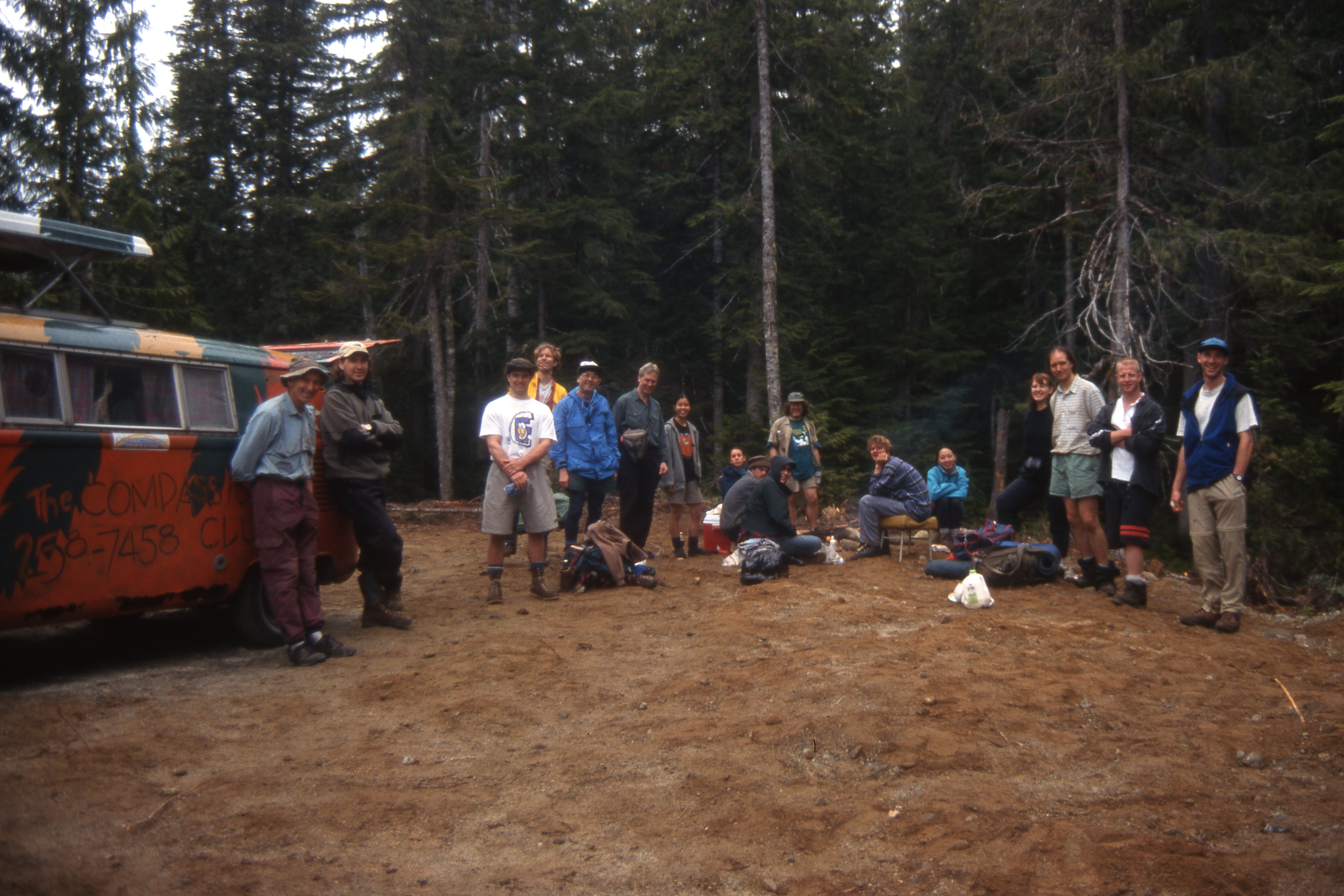 A group of people gathered in a dirt clearing, with an orange Volkswagen van on the left and forest in the background. This is at the Elaho trail head.