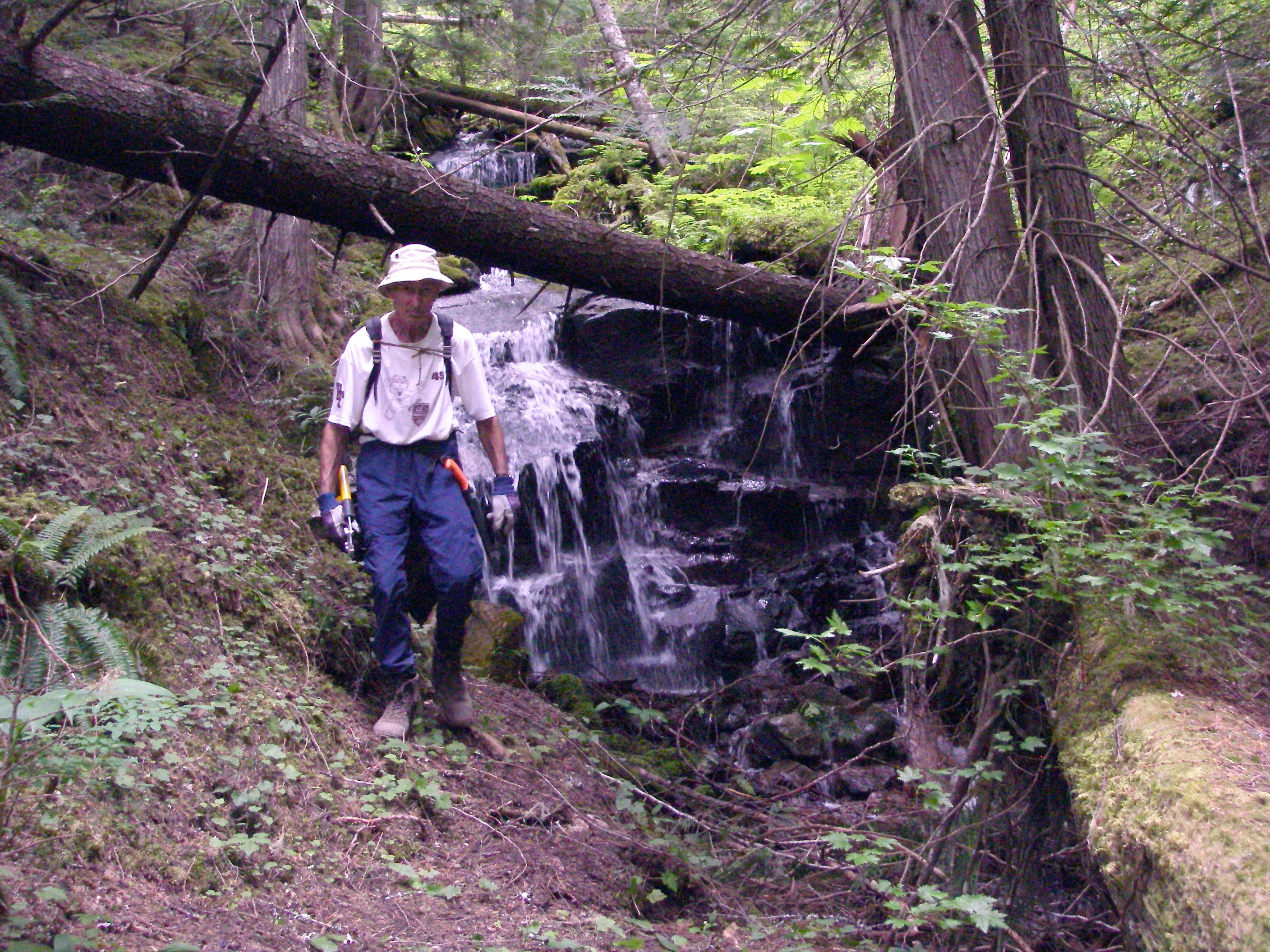 Don Gillespie with his backpack on and clippers in hand in the forest next to a small waterfall.