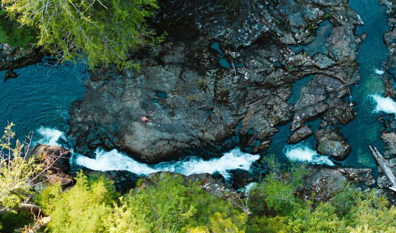 An aerial shot of a river in the Walbran valley, There are large rocks, and a person is standing over them, touching the water. End of image description,