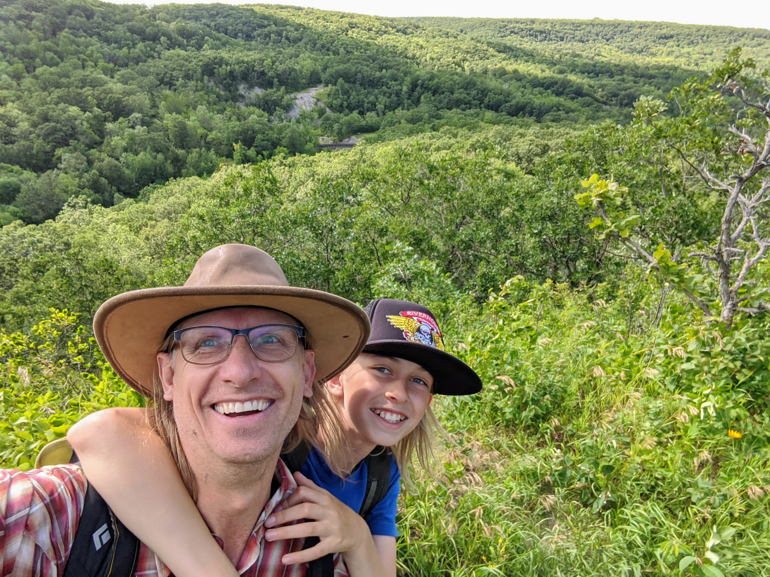 A young boy and his father smile for the camera as a valley spreads below them.