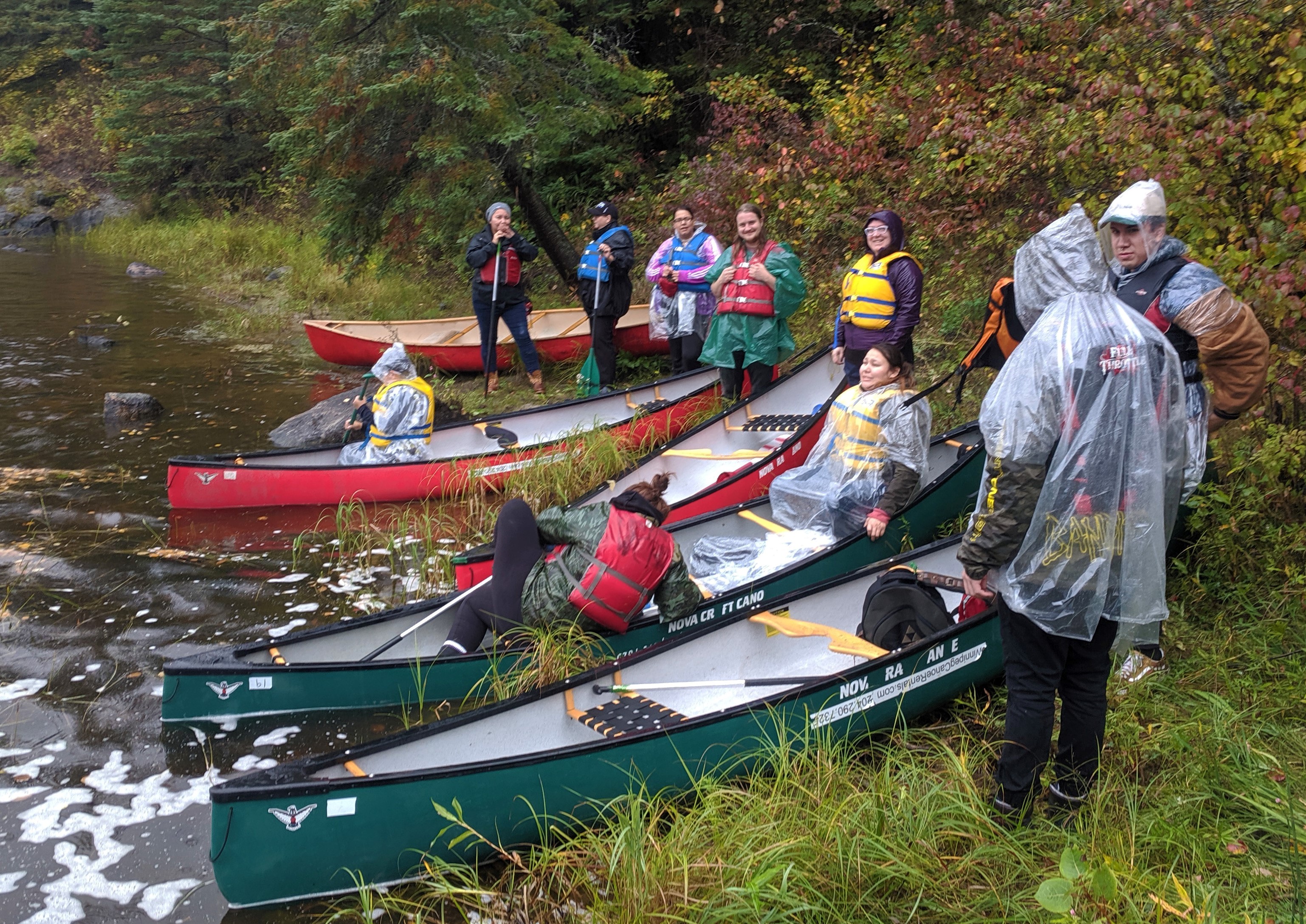 People in colorful rain jackets launch canoes on a lush green bank.