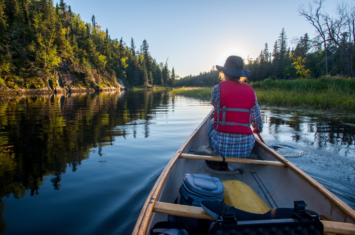 Photo from the back of a red canoe of a person paddling across a lake into the sun.