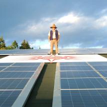 A T'sou-ke person standing above solar panels on a roof.