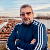 A person wearing glasses and a blue jacket posing in front of logging mill. End of image description.