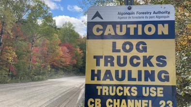 A sign next to a gravel logging road reads "Caution. Log trucks hauling. Trucks use CB Channel 23".