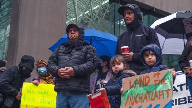 Members of Nuchatlaht Nation stand in the rain holding brightly coloured signs that say "WE WERE TORN DOWN" and "LAND OF THE NUCHATLAHT". End of image description.