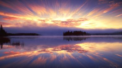 A bright sunburst streaks out from behind pink clouds on a periwinkle sky. Across the lake on a distant shore the forests are silhouetted in black. The water of the lake reflects the sky like a mirror.
