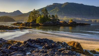 A small lump of lands with fir trees growing sticks out from the shallow shoreline of a lake with smooth waters. A towering mountain covered in forest rises in the background.