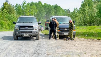 Large, dark-colored trucks sit on the shoulder of a gravel road. A Machine gun is on the ground in front of them. Three men surround it, two are in camo uniforms while one is in all black.