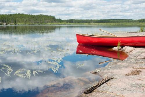 A red canoe on the rocky shore of a big lake, with forested shores and cloudy prairie sky in the background