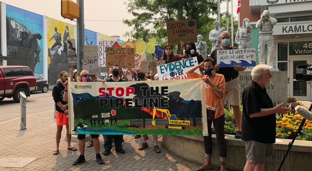 A group of people hold a "Stop the Pipeline" banner and other signs below a smoky sky in front of a local government office in Kamloops.