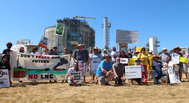 A crowd of people gathered on Tilbury LNG's lawn with signs and banners like "Don't frack our future, no LNG." A gas storage tank is in the background.