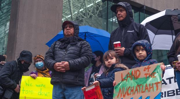 Members of Nuchatlaht Nation stand in the rain holding brightly coloured signs that say "WE WERE TORN DOWN" and "LAND OF THE NUCHATLAHT". End of image description.