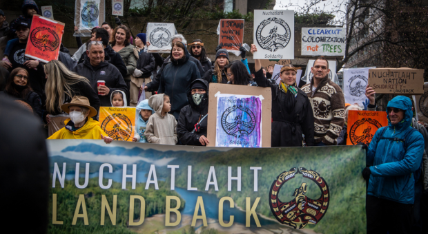 A group of people holding up various signs and banners expressing support for the Nuchatlaht Nation. End of image description. 