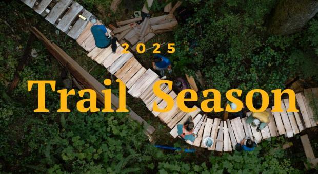 An aerial shot of people working on a wooden trail in the middle of the forest. Text on the image says "2025 Trail Season." 