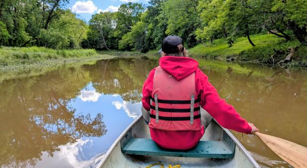 A young man in a red shirt and life jacket, his blond hair sticking out from under a black cap, sits at the front of a canoe facing away from the camera. He paddles through the glossy brown water of the Seine river in Winnipeg, framed by thick green grasses and the hanging boughs of healthy trees.