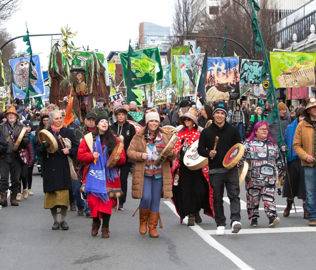 A group of people marching down a street. Some people are holding drums and others are holding banners.