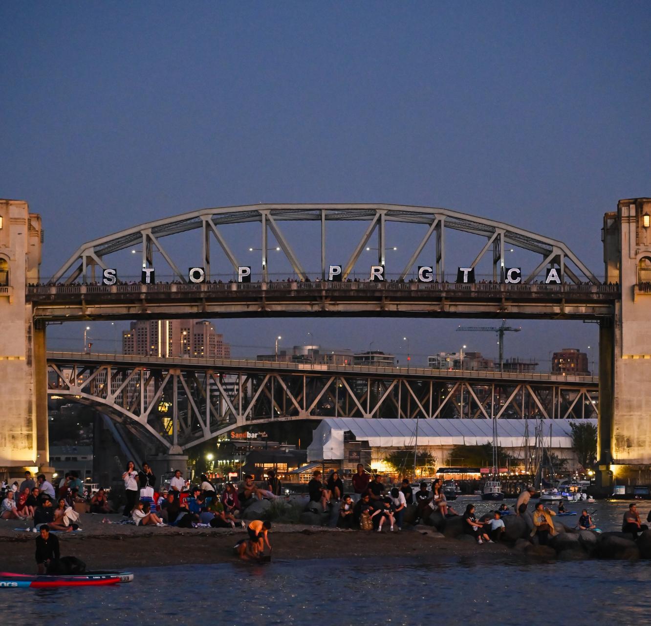 A call to stop PRGTCA in lights shine on a bridge against a dusky summer evening sky
