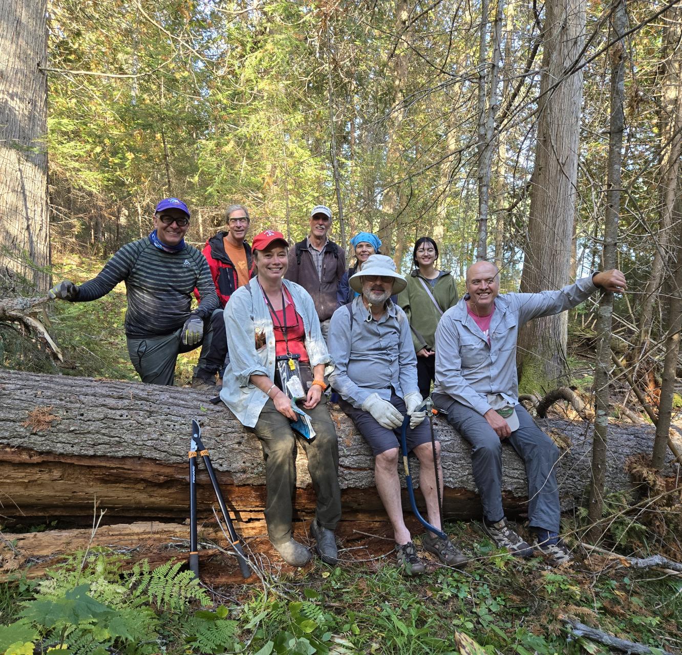 A group of people in Tamegami forest, sitting on a log, holding up gardening equipment, and smiling at the camera. End of image description.