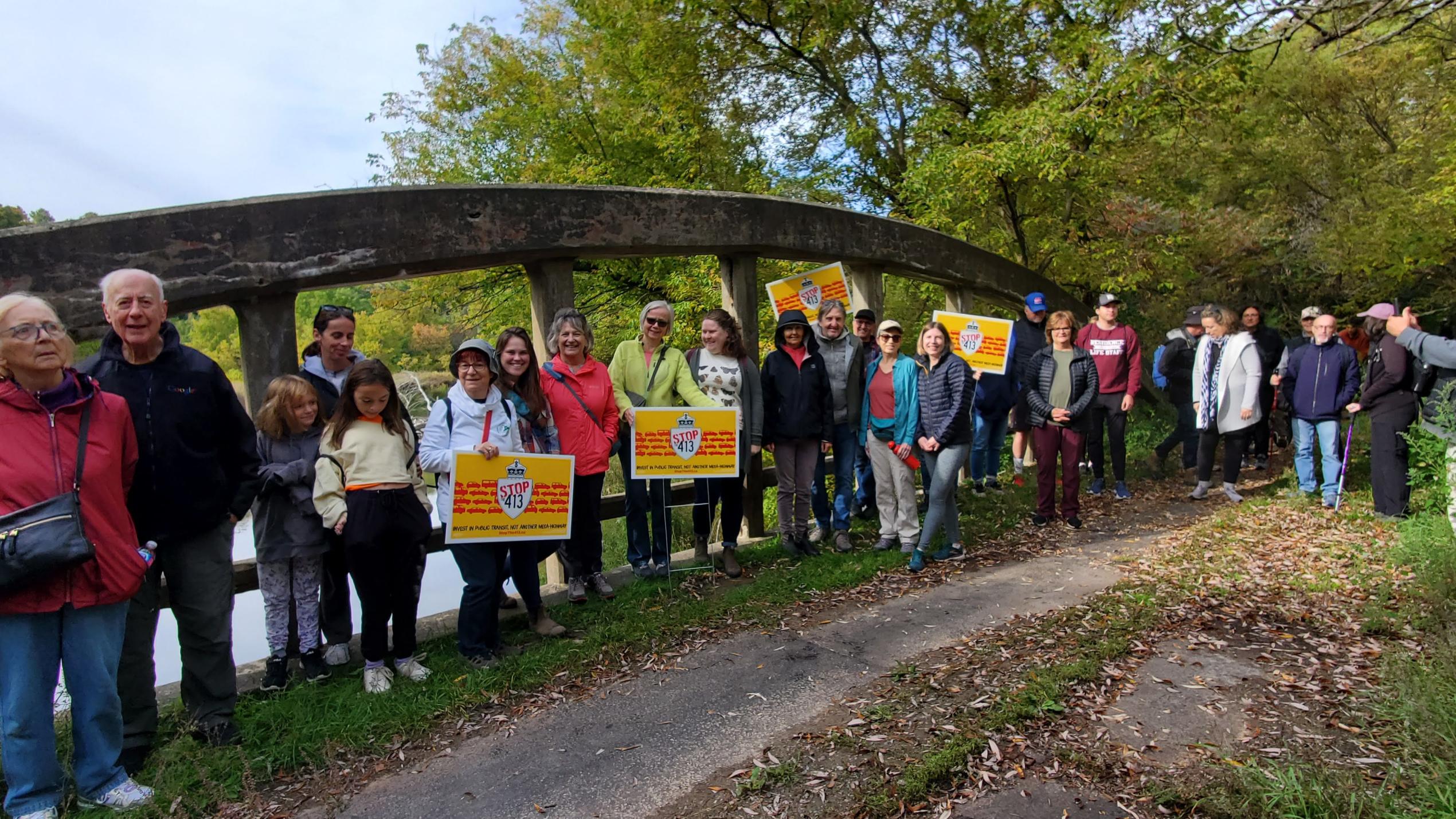 a long line of people stand next to a dirt trail, many of them holding right yellow signs that say "Stop Highway 413"