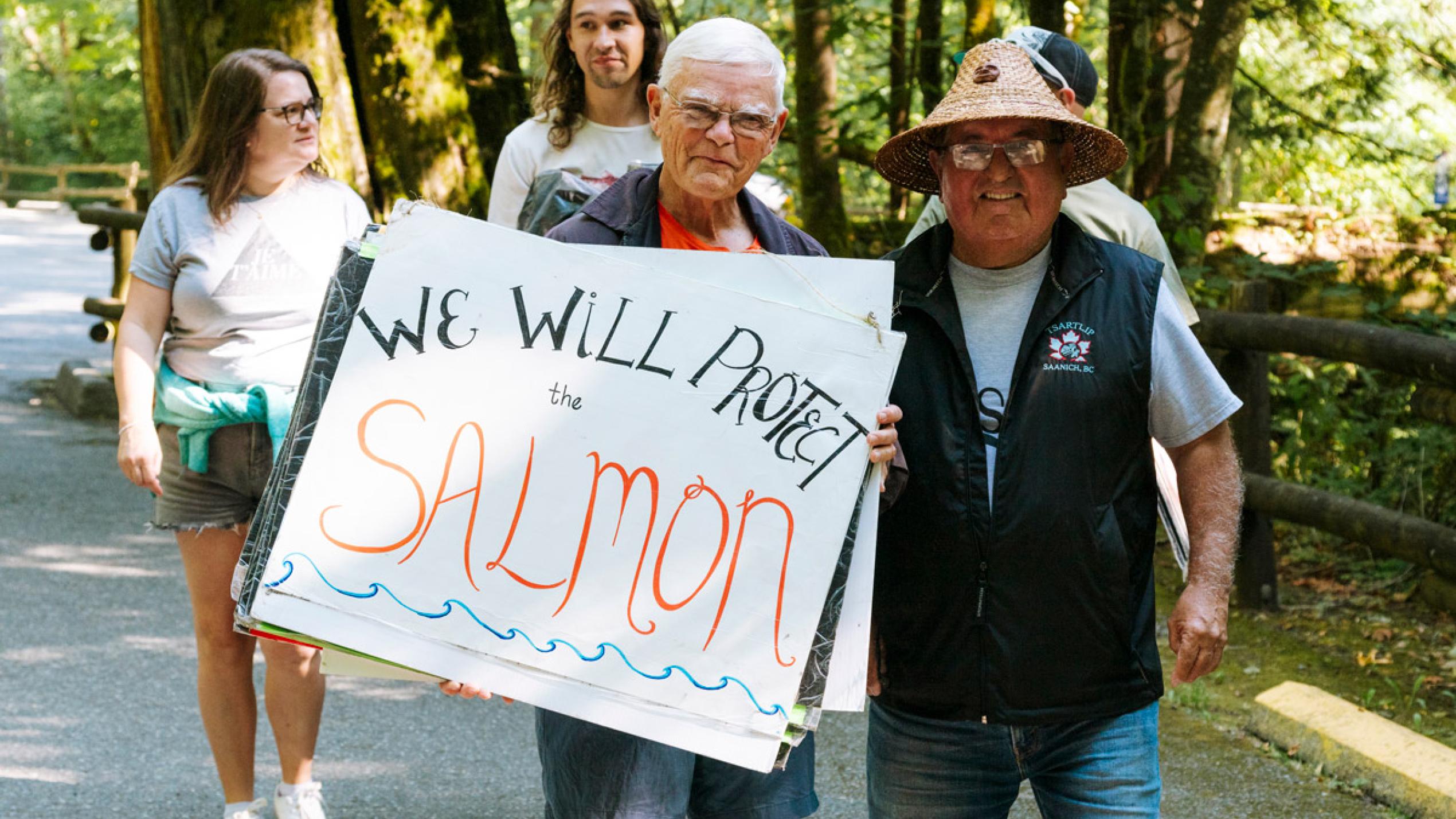 A small group of people stand around a home-made sign reading "We Will Protect the salmon" 