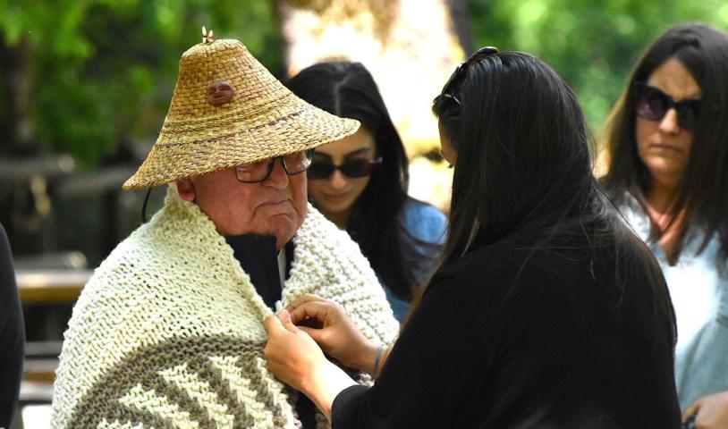 A person is wrapping Elder ZȺWIZUT (Carl Olsen) in a blanket. Elder ZȺWIZUT is wearing a weaved hat. End of image description.