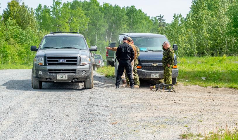 Large, dark-colored trucks sit on the shoulder of a gravel road. A Machine gun is on the ground in front of them. Three men surround it, two are in camo uniforms while one is in all black.