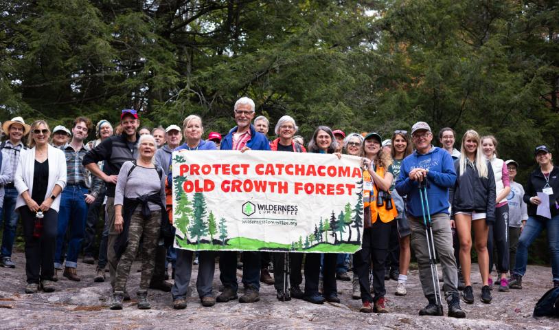 A large crowd to people in hiking gear smile for the camera. In front of them is a hand-painted banner that says "Protect Catchacoma Old-Growth Forest"