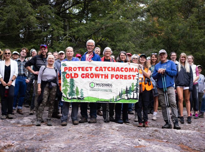 A large crowd to people in hiking gear smile for the camera. In front of them is a hand-painted banner that says "Protect Catchacoma Old-Growth Forest"